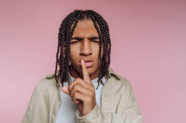 Genç bir adam pembe bir zemine karşı duruyor, parmağını dudaklarına kaldırıyor. Örülmüş saçı benzersiz tarzına renk katıyor. Gizlilik ya da sükunet duygusunu ifade ediyor..