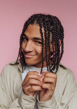 Genç bir adam gülümser ve ellerini şakacı bir şekilde bir arada tutar. Saç stili düzgünce bükülmüş ipliklerden oluşuyor. Pembe bir arka planı olan modern bir kapalı alanda..