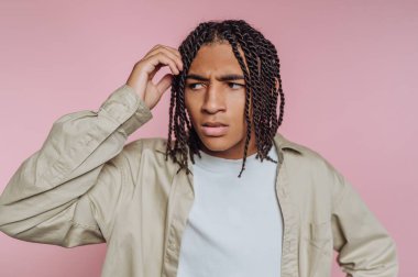 Genç bir birey düşünceli bir ifadeyle ayakta duruyor, başını kaşıyarak. Arkaplan yumuşak bir açık pembe, canlı ve neşeli bir atmosfer yaratıyor..