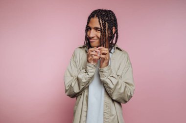 Genç bir adam, ellerini şakacı bir şekilde yüzüne yakın tutarken sırıtarak, yumuşak pembe bir zemin üzerinde duruyor. Örgülü saçı ve rahat gömleği neşeli ve tasasız bir ruh halini yansıtıyor..