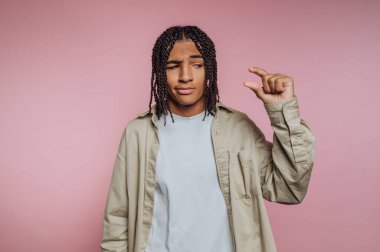 Genç bir insan pembe bir zemine karşı duruyor, parmaklarıyla küçük bir beden göstermek için şakacı hareketler yapıyor. Açık renkli gömlek ve bej ceket giyiyorlar..