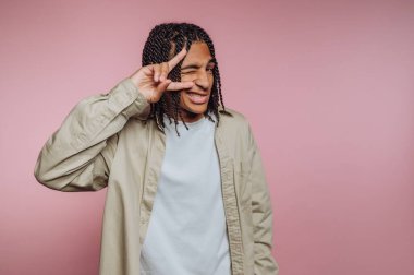 Genç bir birey, yumuşak pembe bir zemine karşı duruyor, gülümsüyor ve sağ eliyle barış işareti hareketi yapıyor. Onların oyuncu tavırları sahneye eğlenceli bir hava katıyor..