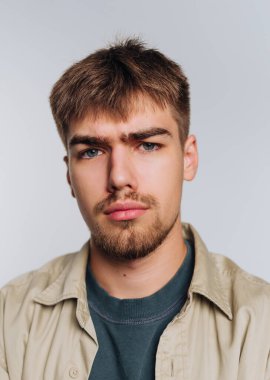 Genç bir adam ciddi bir ifadeyle özgüvenle ayakta duruyor. Onun kısa, stil sahibi saçları ve açık renk bir sakalı var. Tişörtün üzerine gündelik bir gömlek giyip rahatlatıcı ama çarpıcı bir görünüm yaratıyor..