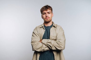 Koyu renk bir tişörtün üzerine açık renk gömlek giyen genç bir adam kollarını kavuşturmuş duruyor. Ciddi ifadesi, basit bir ortamda güven ve kararlılık olduğunu gösteriyor..