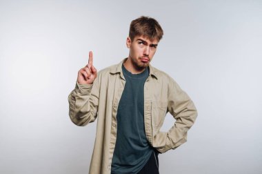 Tarzlı saçlı genç bir adam sade bir arka planda güvenle durur. Bir parmağını kaldırıyor, neşeli bir ifade sergiliyor, tişört ve tişört giyiyor..