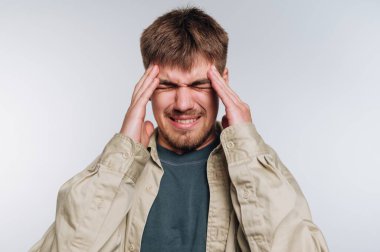 Genç bir adam iki eliyle başını tutuyor, baş ağrısından rahatsızlık duyuyor. Acı dolu bir ifadesi var. Basit bir iç mekan ortamında stres ve yorgunluğu gösteriyor..