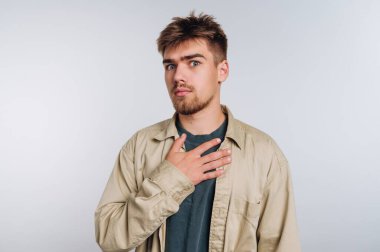 Genç bir adam minimalist bir stüdyoda duruyor, eli kalbine yerleştirilmiş şaşırmış bir şekilde bakıyor. Kısa saçları ve şaşkınlığın nötr bir ifadesi var..