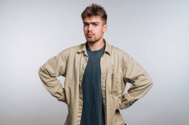 Genç bir adam elleri kalçasında duruyor, ciddi bir ifade sergiliyor. Koyu renk bir tişörtün üzerine bej bir gömlek giyiyor. Stüdyoda açık gri bir arka planda geçiyor..