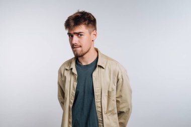 Genç bir adam ellerini arkasina koyup rahat bir poz verir. Kısa saçlı ve sakallı, tişörtünün üzerine açık renkli bir ceket giymiş..