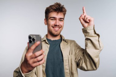 Genç bir adam bir elinde akıllı bir telefonla ayakta duruyor ve neşeli görünerek parmağını havaya kaldırıyor. Parlak ışıklandırma bu kutlama anı için olumlu bir atmosfer yaratır..