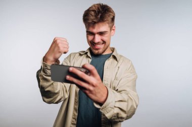 Genç bir adam akıllı telefonunu tutarken sevinçle gülümsüyor, kutlama için yumruğunu sallıyor. Parlak ışıklandırma, oyun zaferi sırasında neşeli bir atmosfer yaratıyor..