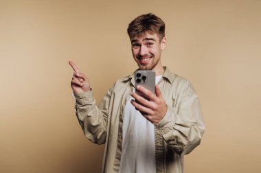 Neşeli genç bir adam düz bir arkaplanda duruyor, bir elinde akıllı bir telefonla gülümsüyor ve işaret ediyor. Rahatlamış ve mutlu görünüyor..