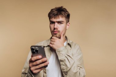 Genç bir adam bir elinde akıllı telefon tutarken önemli bir şey düşünüyor. Düşünceli bir ifadesi var ve sıradan giyinmiş, sade bir arkaplanın önünde duruyor..
