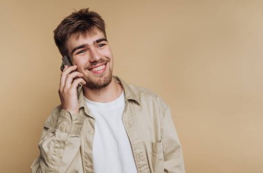 Dostça gülümseyen genç bir adam kulağına telefon dayıyor. Sıcak bej bir duvarın önünde dururken gevşemiş ve sohbete dalmış görünüyor. Güneş ışığı nazikçe sahneyi aydınlatır..