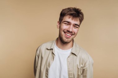 Kısa saçlı ve açık renk gömlekli genç bir adam bej bir arka planda dururken parlak bir şekilde gülümsüyor. İfadesi mutluluk ve sıcaklık yayarak dostça bir hava katıyor..