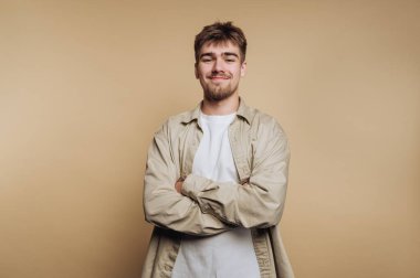 Kolları bağlı genç bir adam, rahatlamış bir güven gösterisi sergiliyor. Beyaz bir tişörtün üzerine hafif bir gülümseme giyer. Arka plan yumuşak, nötr bir ton..