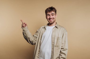 Açık kahverengi saçlı genç bir adam, gülümserken ve yan tarafı gösterirken bej bir gömlek ve beyaz bir tişört giyer. Tarafsız renkli bir arka plana karşı duruyor, neşeli bir hava yaratıyor..