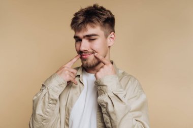 Genç bir adam düz bir arkaplana karşı duruyor, yüzünde küstah bir ifadeyle gülüşünü şakayla işaret ediyor. Neşeli ve neşeli görünüyor..