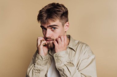 Tarafsız bir arkaplanın önünde duran genç bir adam derin düşüncelere dalmış bir ifadeyle sakalına hafifçe dokunuyor. Açık renkli bir gömlek giyiyor, rahat bir hava veriyor..