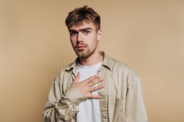 Genç bir adam şaşırmış bir ifadeyle duruyor, bir elini göğsüne koyuyor. Günlük kıyafetleri basit bej arka planla çelişiyor ve duygusal bir ortam yaratıyor..