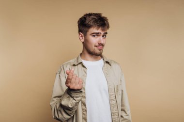 Genç bir adam gülümsüyor ve bronzlaşmış bir arka plana karşı neşeli bir el hareketi yapıyor. Açık renkli bir gömlek giyiyor ve yüzünde kendinden emin bir ifade var..