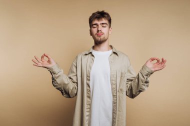 Genç bir adam elleri mudra pozisyonunda ve gözleri kapalı meditasyon yapar. Açık renkli bir odada duruyor, huzur ve sükunet hissi veriyor..