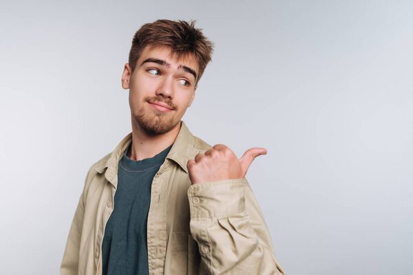 Молодой человек стоит с уверенной улыбкой, небрежно указывая на себя. Он носит светлую рубашку поверх тёмной футболки, создавая расслабленный вид. Его игривое выражение лица добавляет очарования моменту.