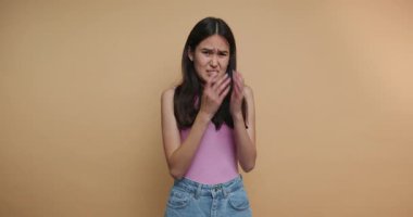 Genç bir kadının görüntüsü gözlerini duygusal yoğunlukla kapatıyor, elleri yüzünü örtüyor, bej arka planda bir anlık hassasiyet yakalıyor.