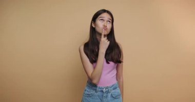 Genç bir kadının görüntüsü düşünceli görünüyor. Parmağı çenesinde ve bej arka planda tuhaf bir ifade var.