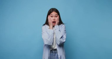 Yüzünde şaşırmış bir ifadeyle genç bir kadının yanaklarını tutarken yaşadığı şoku gösteren görüntüler. Mavi arka planda sıradan bir kıyafet giyiyor.