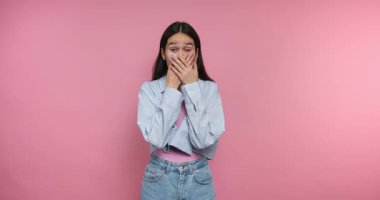 Genç, şaşkın bir kadının, pembe arka planda şaşkınlık ve şoku yansıtırken ağzını şaşkınlıkla kapadığı görüntüler.