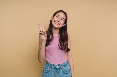 Bej arka planda barış işareti gösteren gülümseyen genç kadın.