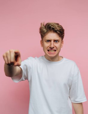 Beyaz tişörtlü genç adam pembe arka planda kızgın bir ifadeyle işaret ediyor..