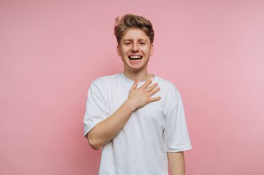 Beyaz tişörtlü genç adam pembe arka planda elleri göğsünde gülümsüyor..