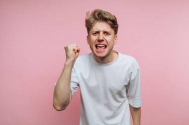 Beyaz tişörtlü genç bir adam pembe arka plana karşı yumruğunu kaldırmış öfkesini ifade ediyor..