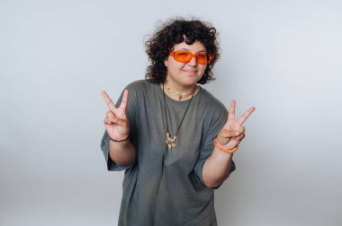 Kıvırcık saçlı, turuncu güneş gözlüğü takan ve barış işareti yapan biri..