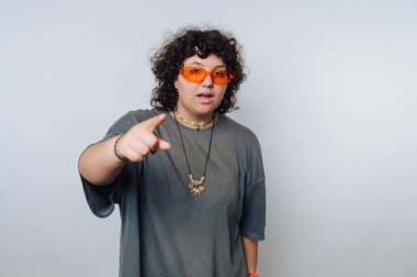 Kıvırcık saçlı, turuncu güneş gözlüğü takan ve öne bakan bir kolyesi olan biri..