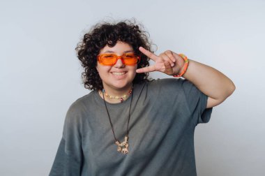 Kıvırcık saçlı, turuncu güneş gözlüklü ve kolyeli gülümseyen biri, arka planda barış işareti yapıyor..
