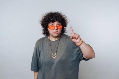 Kıvırcık saçlı, turuncu güneş gözlüklü, gri gömlekli, bir parmağını kaldıran..