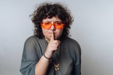 Kıvırcık saçlı, turuncu güneş gözlüğü takan biri susma hareketi yapıyor..