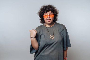 Kıvırcık saçlı, turuncu güneş gözlüklü ve gri tişörtlü, başparmağı omzunda..