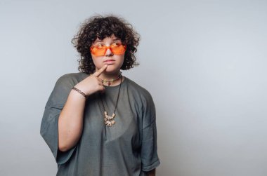 Kıvırcık saçlı, turuncu güneş gözlüklü ve gri gömlekli biri, özenle arka planda poz veriyor..