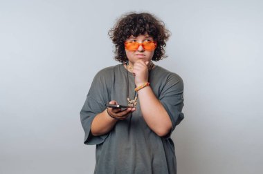 Kıvırcık saçlı, turuncu güneş gözlüğü takan, elinde telefon tutan ve arka planda düşünceli görünen biri..