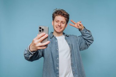 Genç adam akıllı telefonuyla selfie çekiyor, gülümsüyor ve mavi arka planda barış işareti yapıyor..