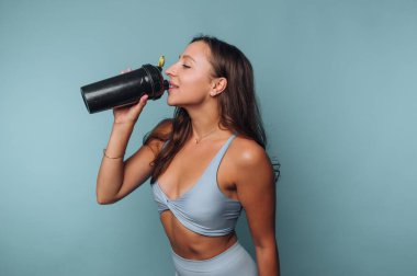 Egzersiz kıyafetli bir kadın mavi arka planda bir şişeden su içiyor..