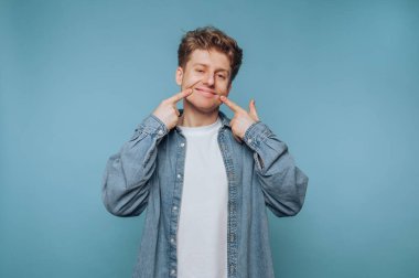 Kot gömlekli genç adam gülümsüyor ve yanaklarını gösteriyor..