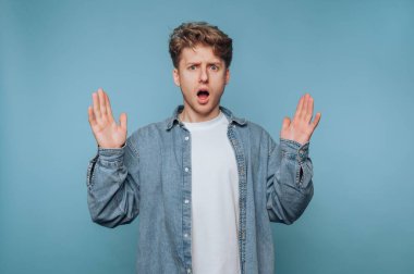 Mavi arka plana el kaldırmış şaşırmış genç bir adam..