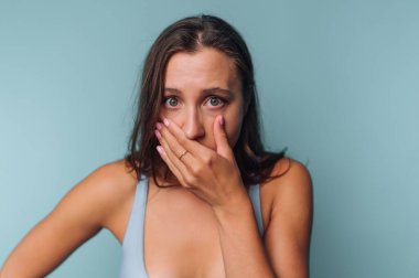 Ağzını mavi arka planda kapatan şaşkın bakışlı bir kadın..