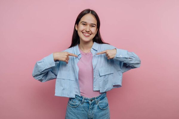 Улыбающаяся молодая женщина в повседневной одежде указывает на себя на розовом фоне.