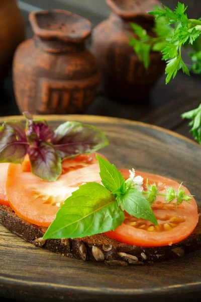 Tmavý chléb bruschetta s červenými rajčaty a zelenými listy bazalky zblízka na dřevěné texturované rustikální desce. — Stock fotografie